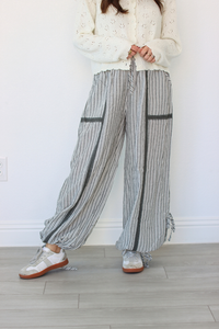 A person wearing loose, wide-leg striped pants with dark vertical stripe panels, a cream knit sweater, and white sneakers, standing against a white wall on a light wood floor.