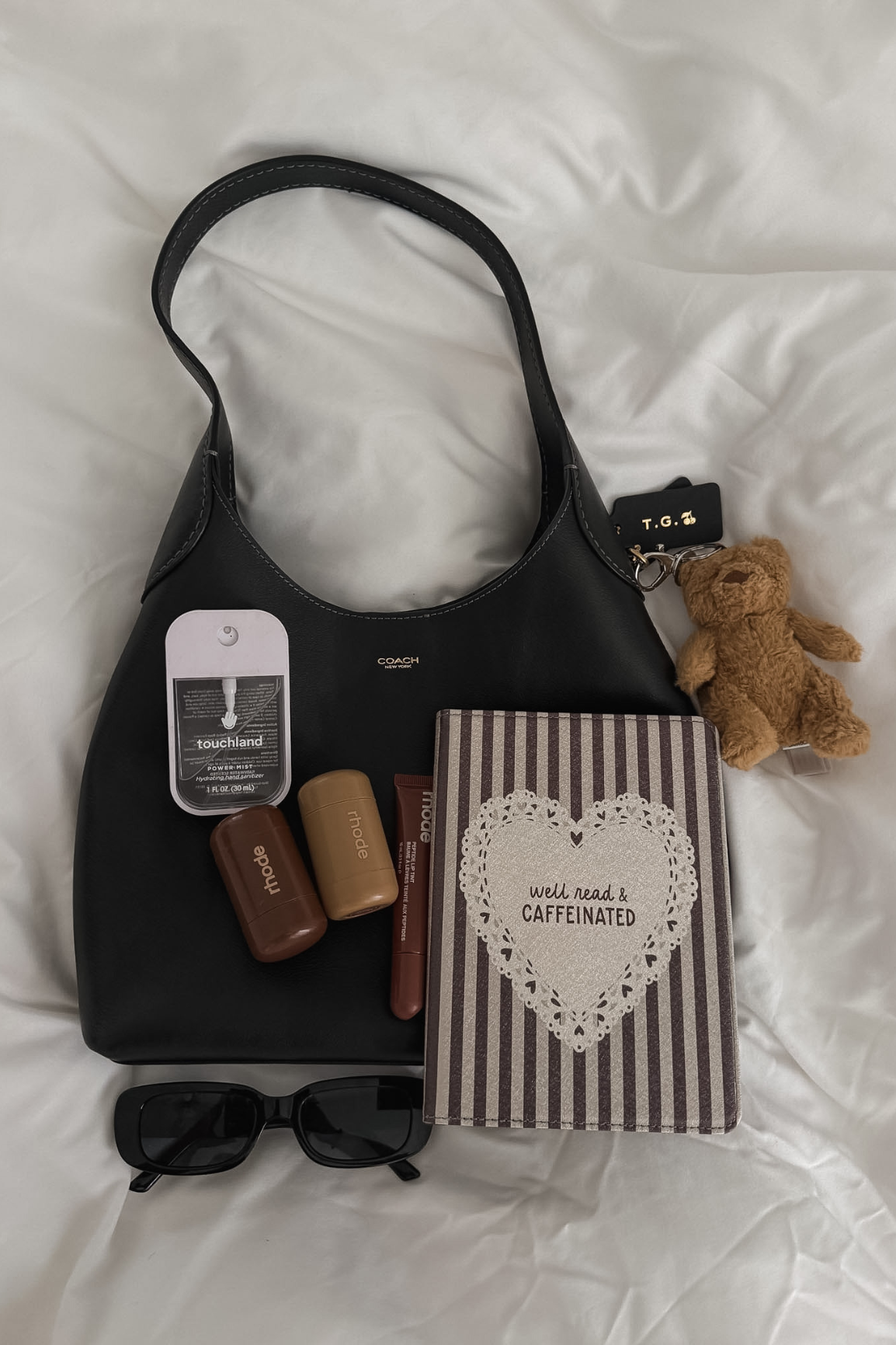 A black shoulder bag laid flat on white bedding with several styled accessories around it, including sunglasses, a striped kindle case, small bottles, a card, and a small teddy bear keychain.