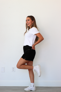 Woman in a white t-shirt, black shorts, and white sneakers posing against a plain wall.