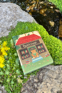 A kindle case with a bookshop design, placed outdoors on mossy rocks beside a stream.