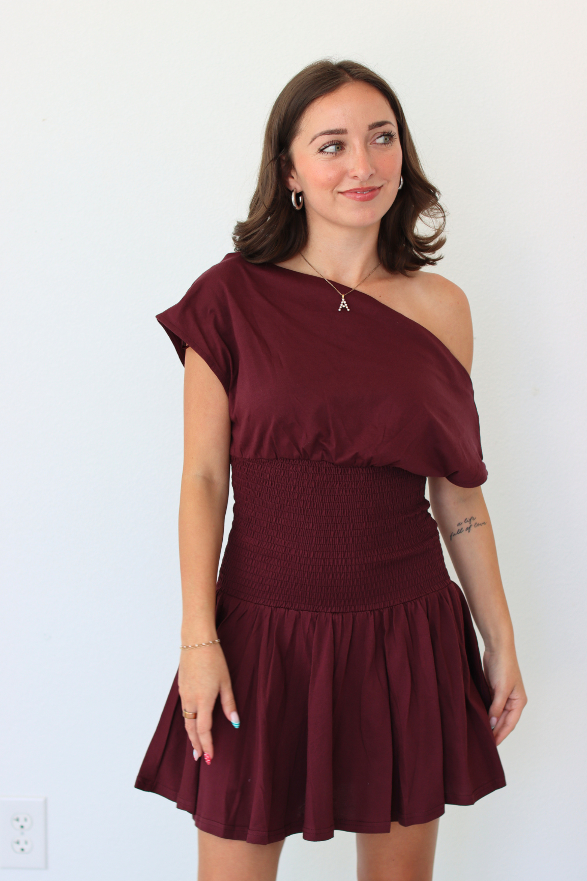 A woman modeling a burgundy one-shoulder mini dress with a smocked waist and flared skirt, standing against a plain white wall.