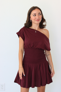 A woman modeling a burgundy one-shoulder mini dress with a smocked waist and flared skirt, standing against a plain white wall.