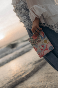 A close-up lifestyle shot of a person holding a kindle case. The person is wearing a white lace-trimmed blouse and blue jeans, standing outdoors against a blurred beach or sunset background.