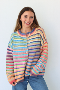 A smiling woman wearing a colorful, fuzzy striped cardigan with blue jeans, standing against a plain white wall.