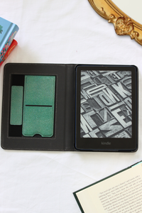 A black Kindle e-reader shown open in a folio-style case on a white fabric surface, with kickstand and hand strap. Decorative books and an ornate gold frame are partially visible around it.