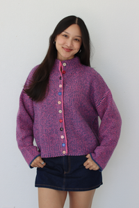A woman wearing a purple and pink marled knit cardigan with colorful buttons, paired with a dark denim skirt, standing against a plain white wall.