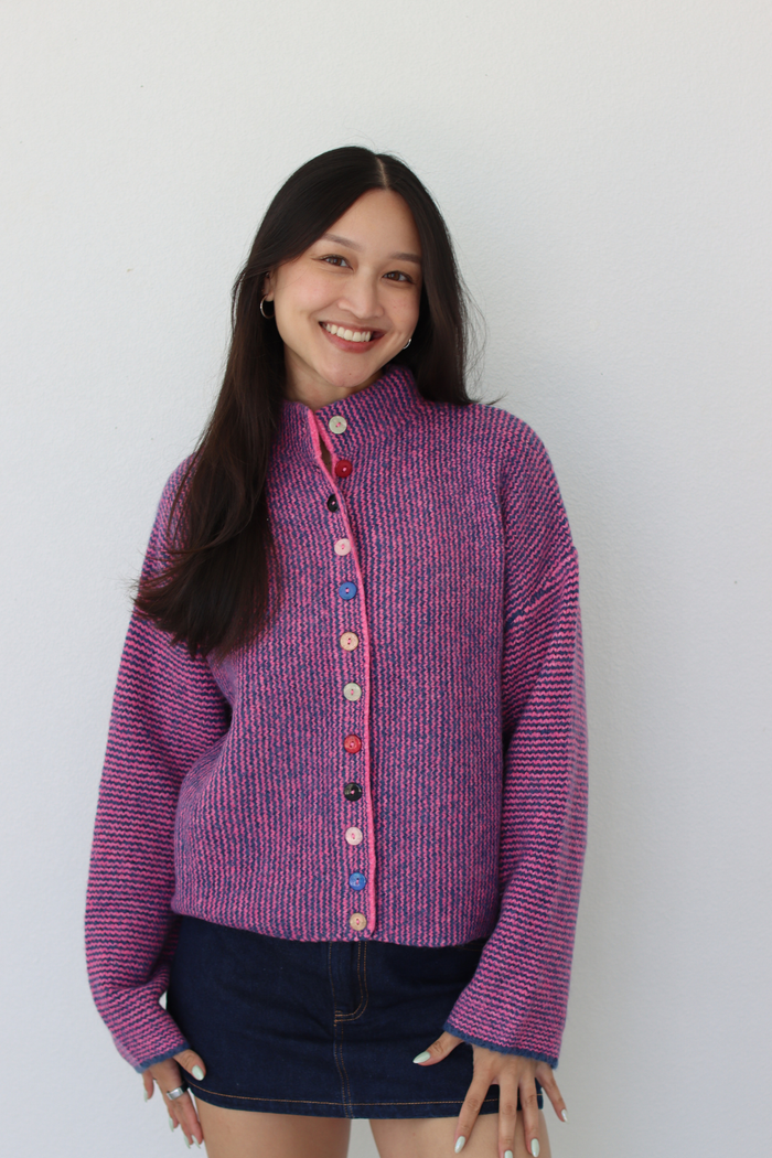 A smiling young woman with long dark hair stands against a plain light gray wall wearing a purple and pink marled knit cardigan with colorful buttons and a dark denim mini skirt.