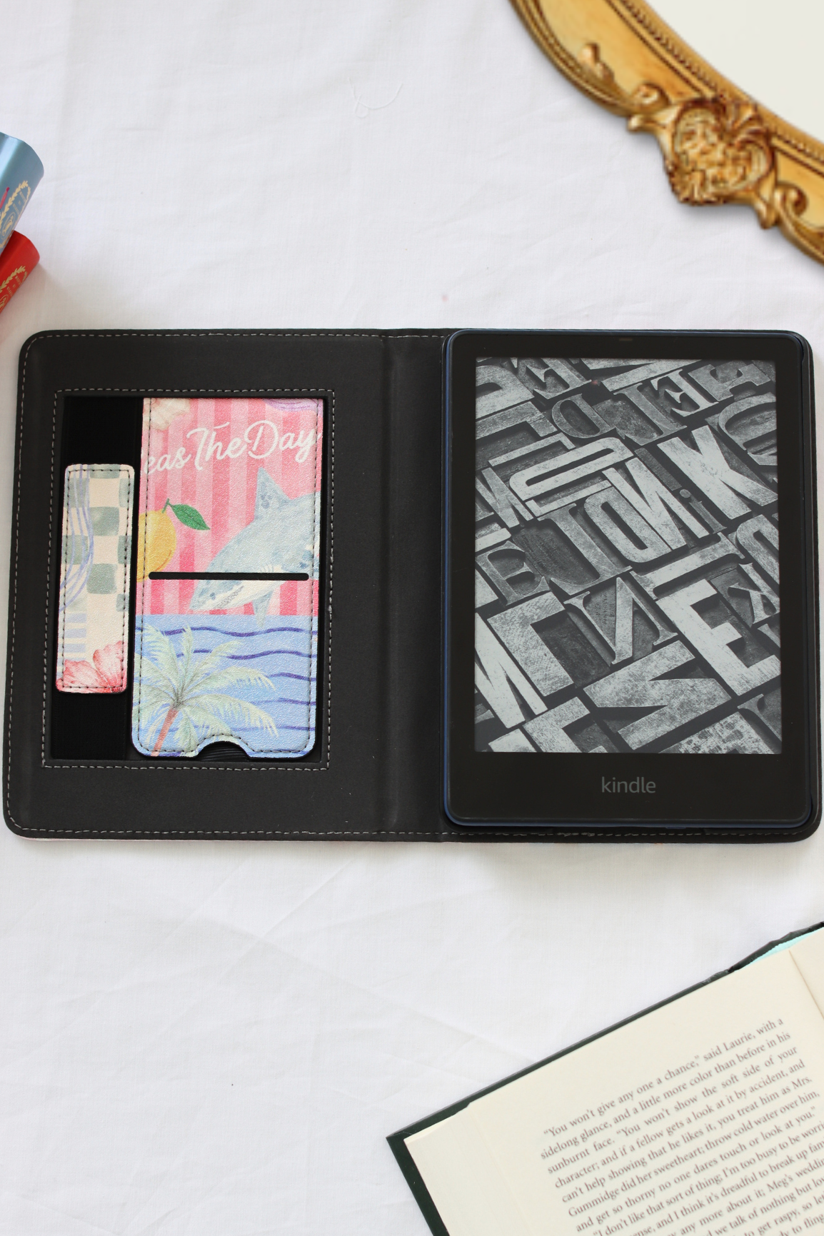 A black Kindle e-reader shown open in a folio-style case on a white fabric surface, with card slots on the left holding colorful cards and an open book in the lower right corner.