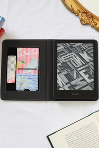 A black Kindle e-reader shown open in a folio-style case on a white fabric surface, with card slots on the left holding colorful cards and an open book in the lower right corner.