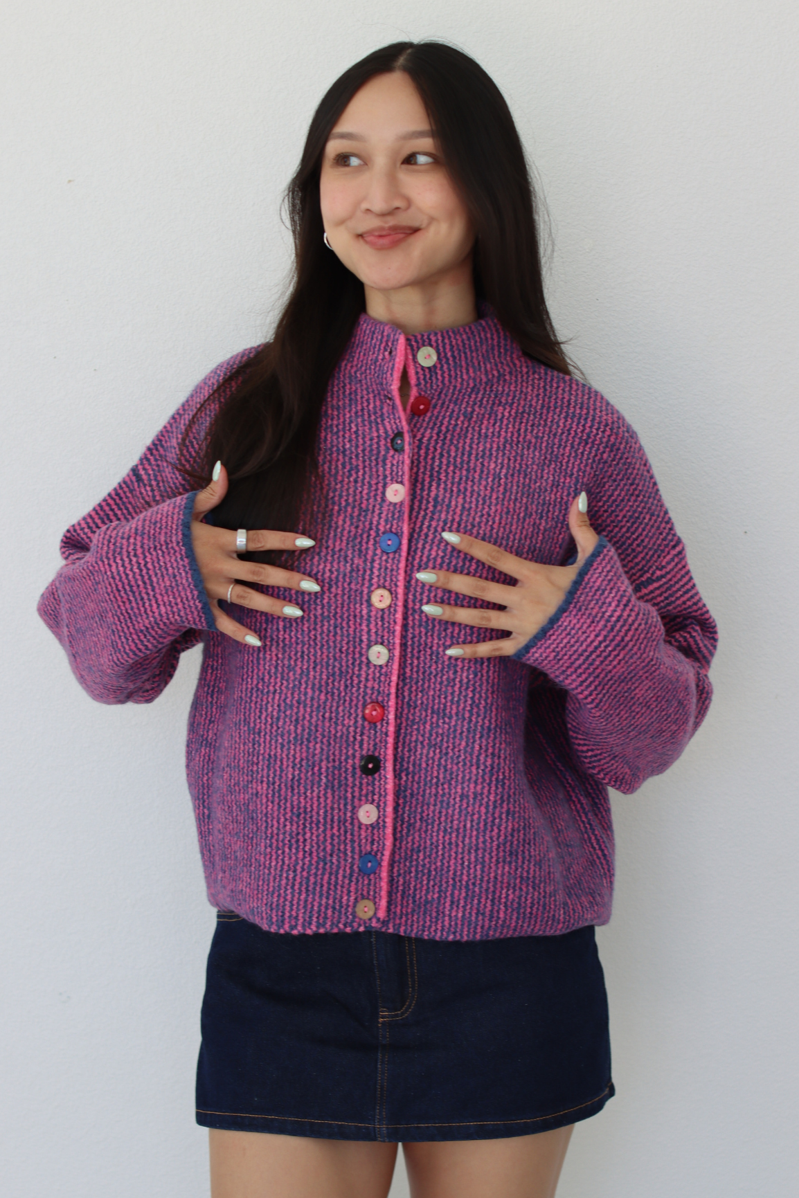 A woman modeling a pink and purple marled knit cardigan with colorful buttons, paired with a dark denim mini skirt, against a plain white wall.