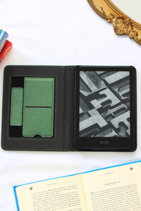Kindle device in a black leather case on a white surface with a book and pen.