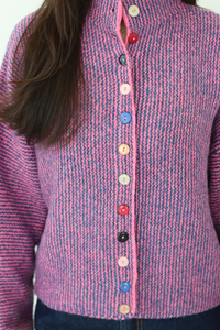 A close-up of a person wearing a pink and purple marled knit cardigan sweater with a high mock neck and colorful decorative buttons down the front.