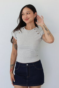 A woman modeling a light gray short-sleeve t-shirt with decorative buttons on the shoulders and a dark blue denim mini skirt, standing against a plain white wall.