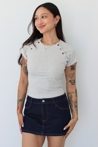 A woman modeling a light gray short-sleeve t-shirt with decorative buttons on the shoulders and a dark blue denim mini skirt, standing against a plain white wall.