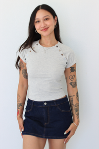 A woman modeling a light gray short-sleeve t-shirt with decorative buttons on the shoulders and a dark blue denim mini skirt, standing against a plain white wall.