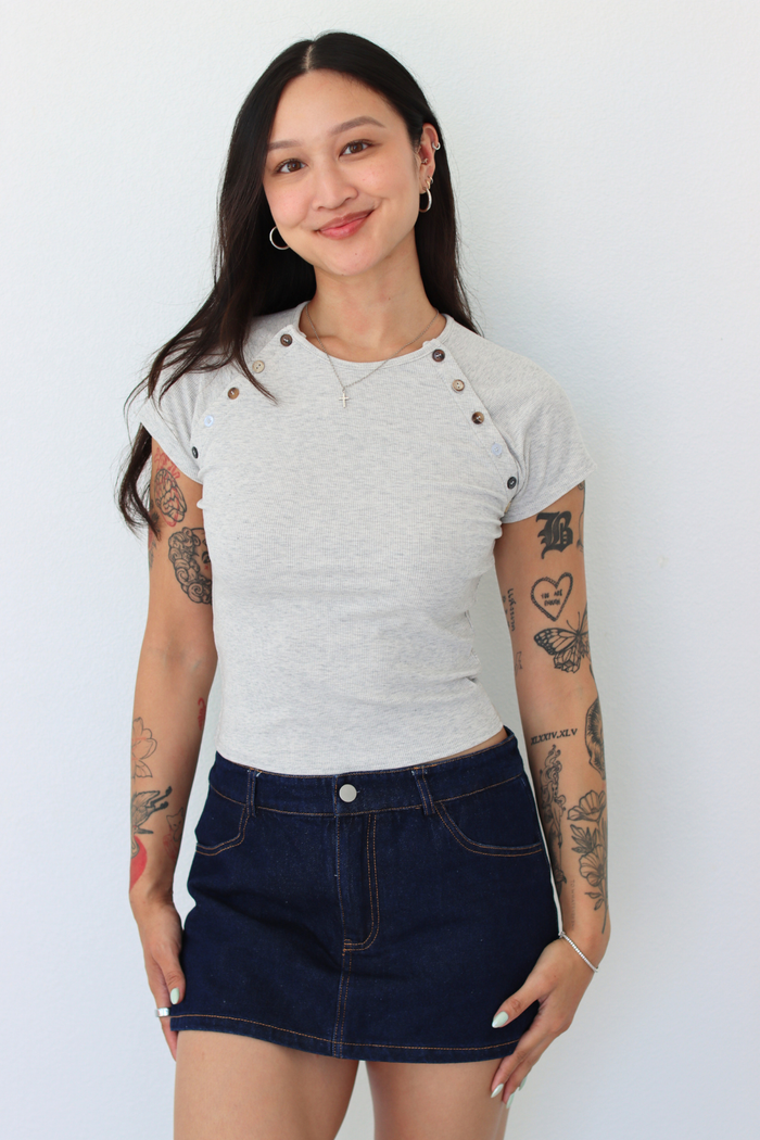 A woman modeling a light gray short-sleeve t-shirt with decorative buttons on the shoulders and a dark blue denim mini skirt, standing against a plain white wall.