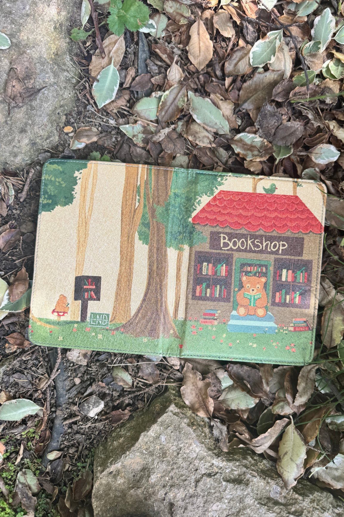 A kindle case lies open on a bed of dry leaves and rocks outdoors. 