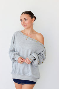 Woman wearing a light gray off the shoulder sweater with buttons along the trim 