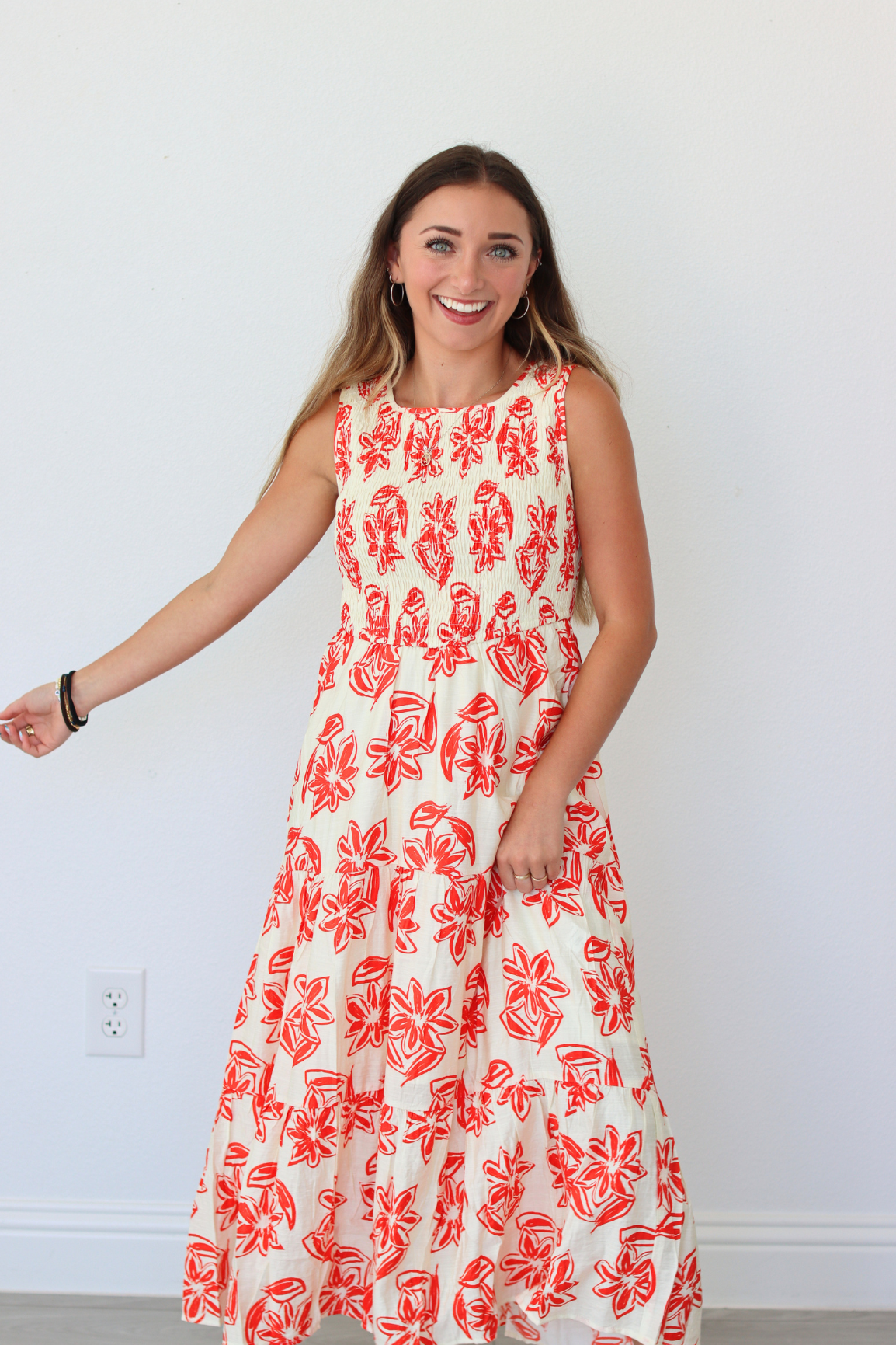 A woman modeling a sleeveless, tiered maxi dress with a red abstract floral print on a cream background, standing against a plain white wall.