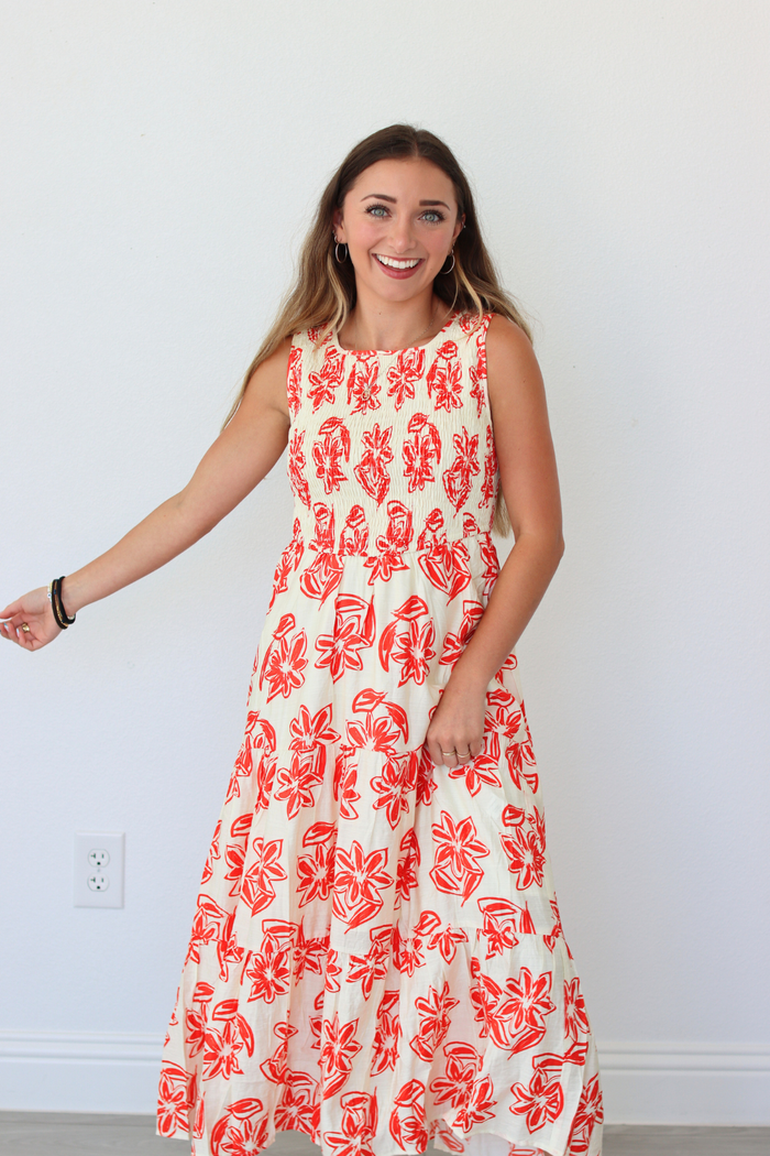 A woman modeling a sleeveless, tiered maxi dress with a red abstract floral print on a cream background, standing against a plain white wall.