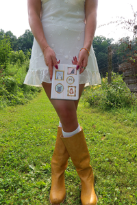 A person outdoors in a white dress and mustard yellow rain boots holds a small white card with several small framed illustrations.