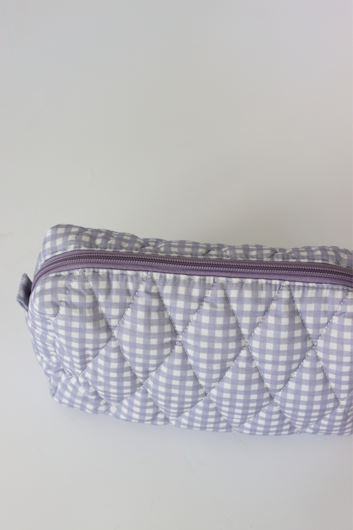 A quilted cosmetic pouch with a light purple and white gingham pattern, featuring a matching purple zipper, shown on a plain light gray background.