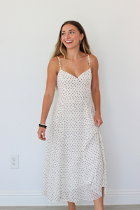 A woman modeling a white spaghetti-strap midi dress with a small black polka dot pattern, standing against a plain white wall.