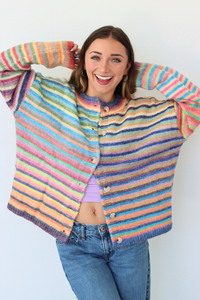 A woman smiling and posing with her arms raised, wearing a colorful striped knit cardigan with pastel rainbow stripes, blue jeans, and a light purple crop top against a plain white background.