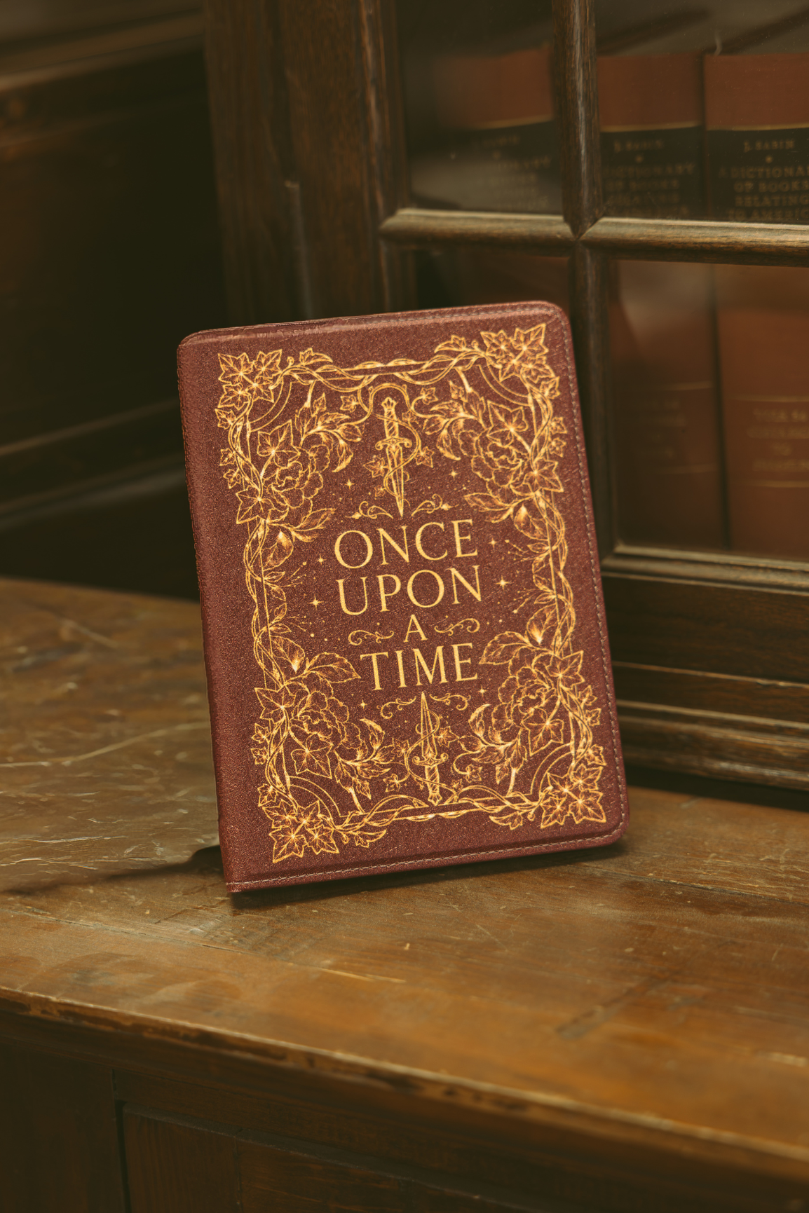 A brown leather-bound book or journal with ornate gold foil detailing and the title 'ONCE UPON A TIME' on the front, standing on a wooden surface with a dark wood bookcase in the background.