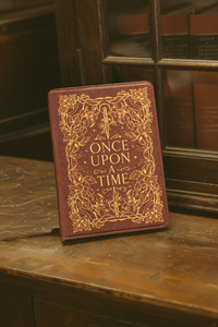 A brown leather-bound book or journal with ornate gold foil detailing and the title 'ONCE UPON A TIME' on the front, standing on a wooden surface with a dark wood bookcase in the background.