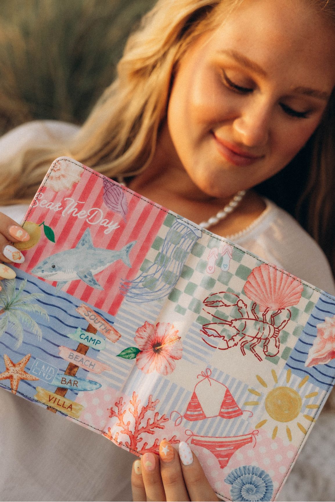 A woman holding a colorful patchwork-style kindle case with nautical and beach-themed illustrations including a lobster, shark, seashell, sun, and striped patterns.