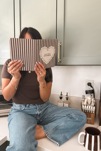 A woman sitting on a kitchen counter holds a striped kindle case in front of her face. The item has brown and white vertical stripes, a white heart-shaped cutout, and the words 'well read & caffeinated' printed inside the heart.