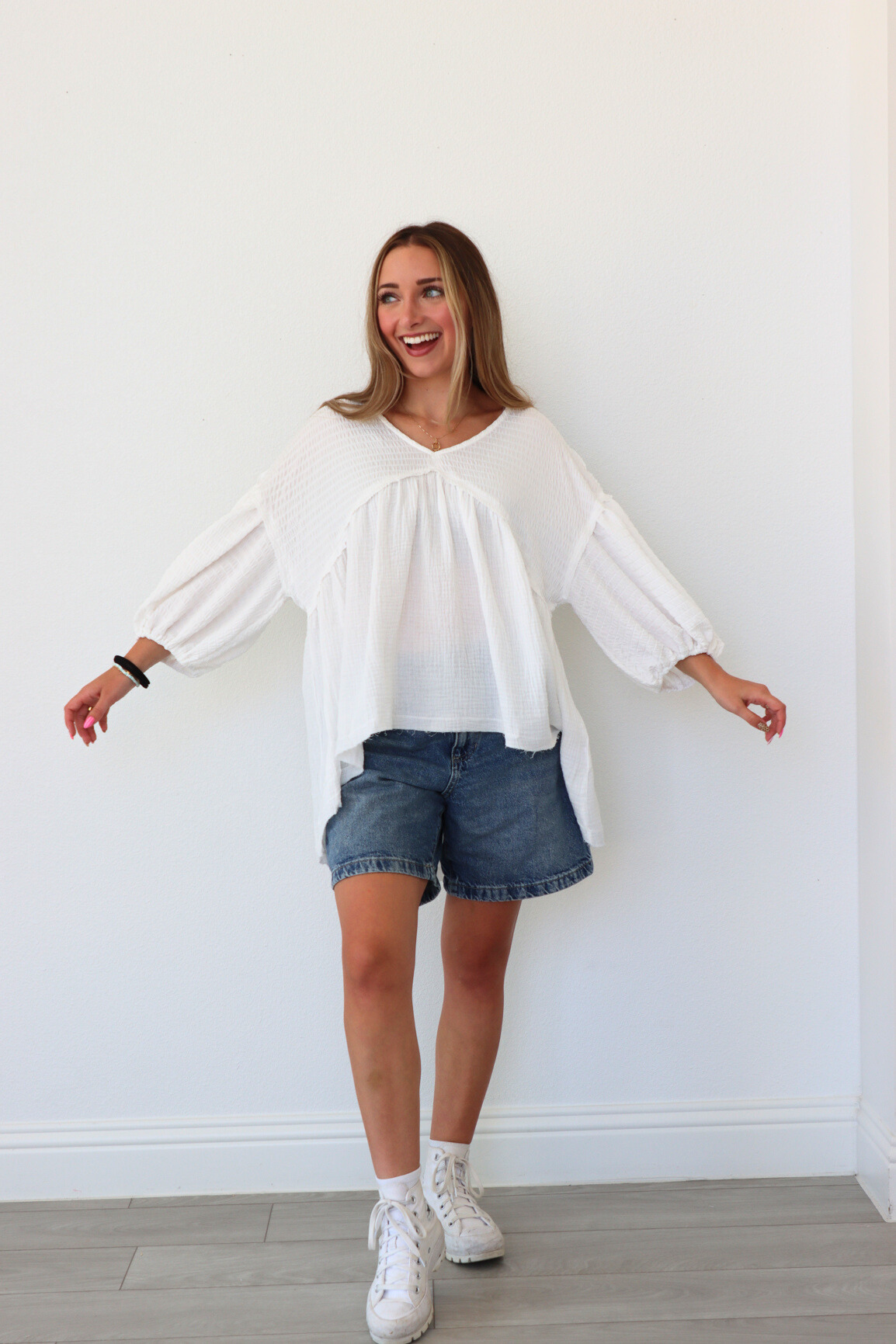 Woman wearing a white blouse, denim shorts, and white sneakers against a white wall.