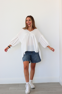 Woman wearing a white blouse, denim shorts, and white sneakers against a white wall.