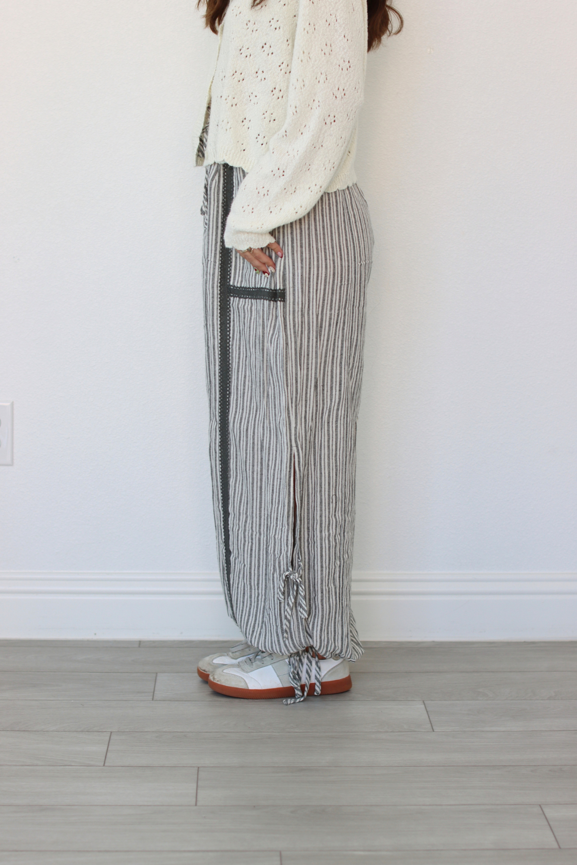 A person wearing loose, wide-leg striped pants with a cream knit top and white sneakers, standing against a plain white wall on a light wood floor.