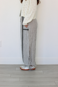 A person wearing loose, wide-leg striped pants with a cream knit top and white sneakers, standing against a plain white wall on a light wood floor.