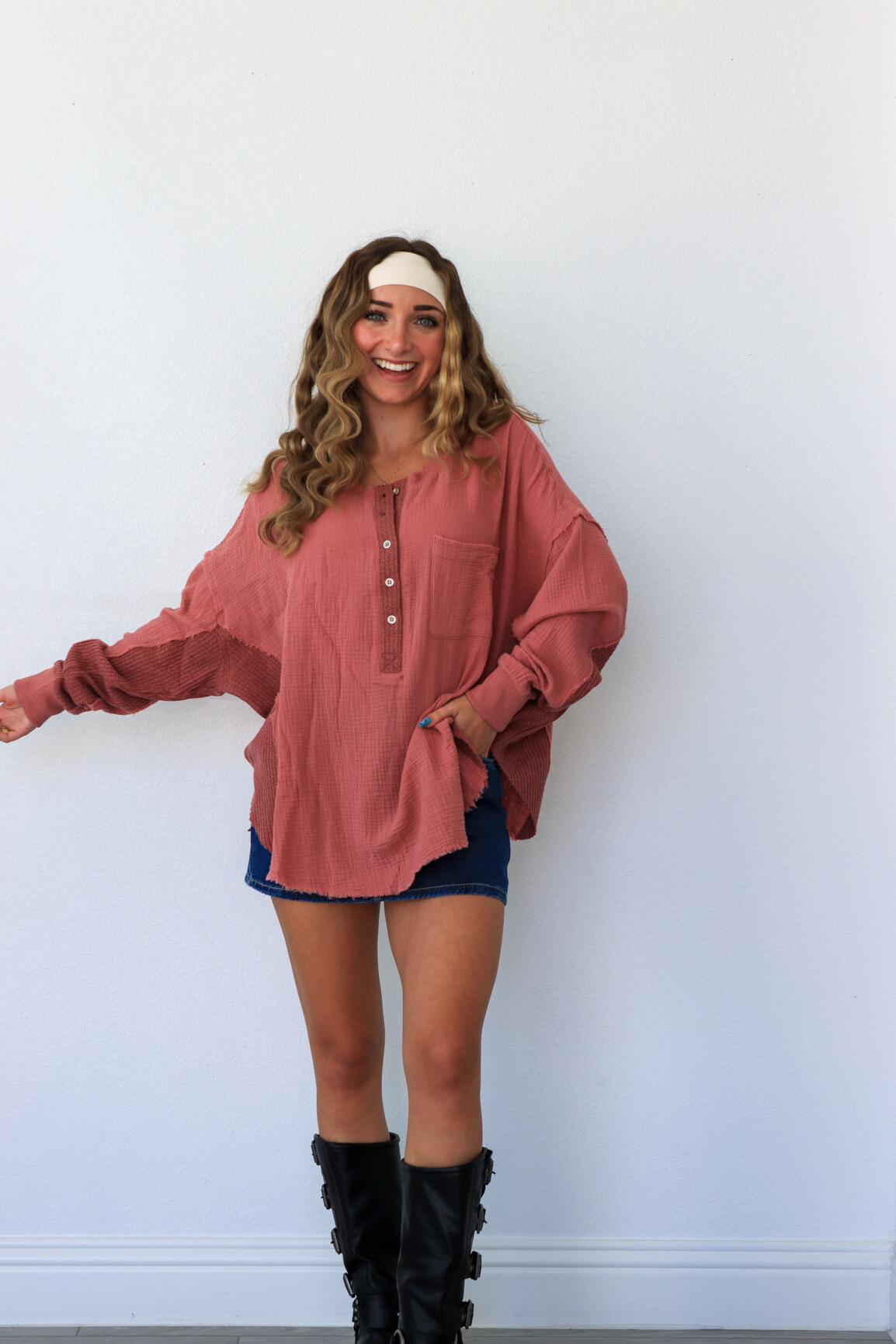 Woman wearing a pink blouse, blue shorts, and black boots against a white wall