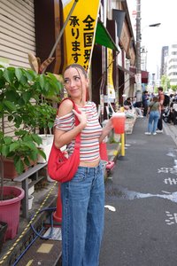 Woman wearing a stripe off the shoulder top holding a red cup on a street with potted plants and a yellow sign in the background
