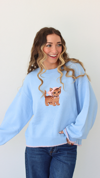 Woman wearing a light blue sweater with a cat embroidery design against a white background 