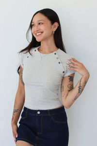 A woman modeling a light gray short-sleeve t-shirt with decorative buttons on the shoulders and a dark blue denim mini skirt, standing against a plain white wall.