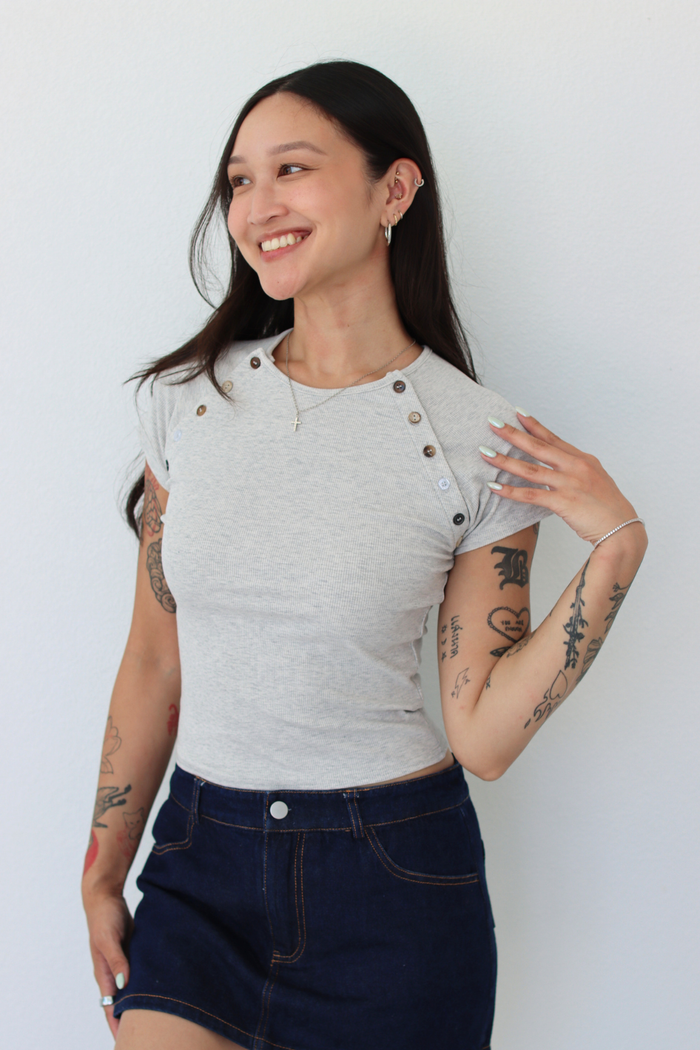 A woman modeling a light gray short-sleeve t-shirt with decorative buttons on the shoulders and a dark blue denim mini skirt, standing against a plain white wall.