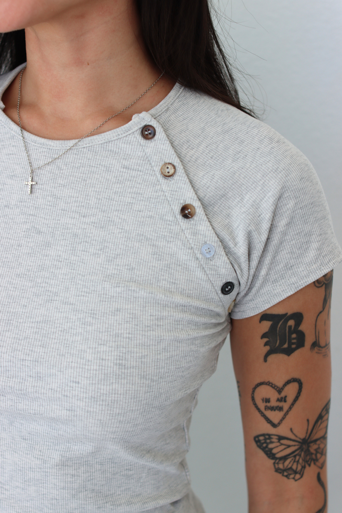 Close-up of a person wearing a light gray ribbed short-sleeve henley shirt with brown buttons down the front placket. The person has visible arm tattoos and a necklace, photographed against a plain light gray background.