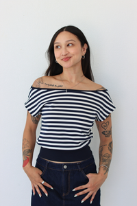 A woman with tattoos wearing a navy and white horizontally striped off-the-shoulder crop top and dark blue jeans, posing against a plain white wall.