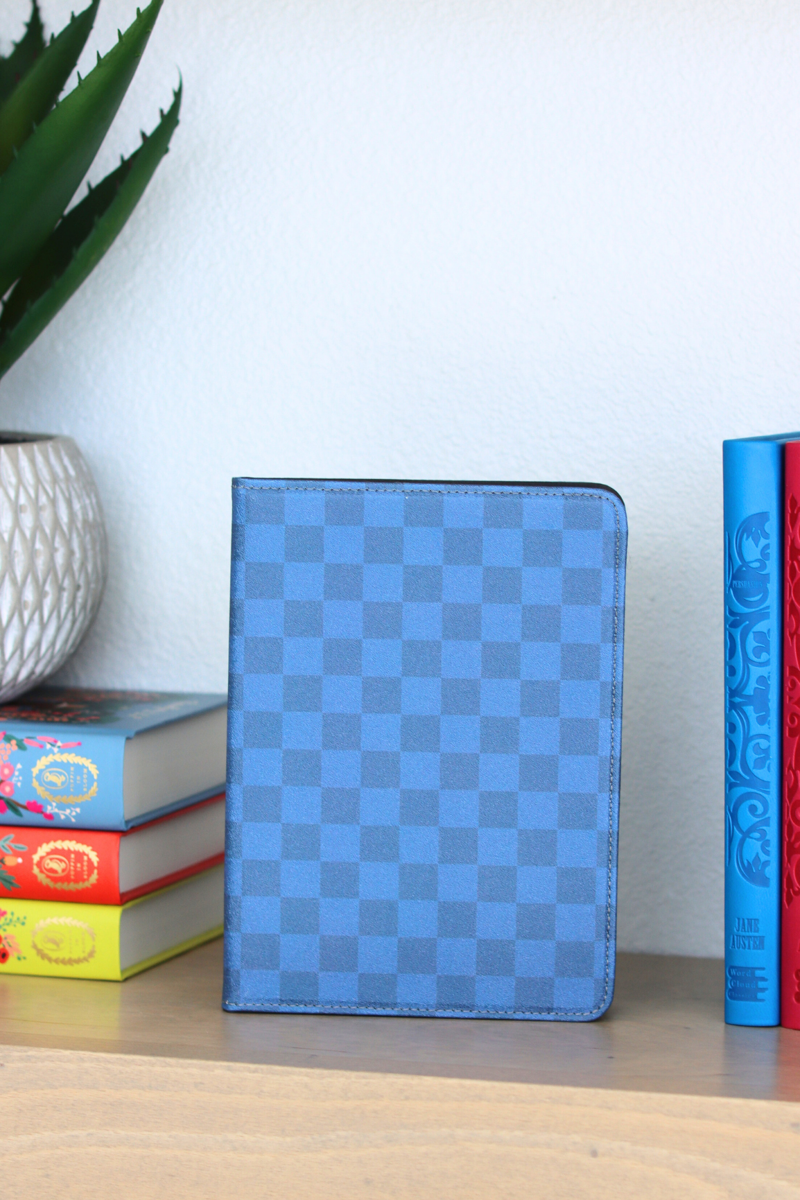 Blue checkered tablet case on a wooden surface with books and a plant in the background