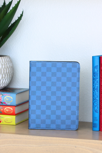 Blue checkered tablet case on a wooden surface with books and a plant in the background