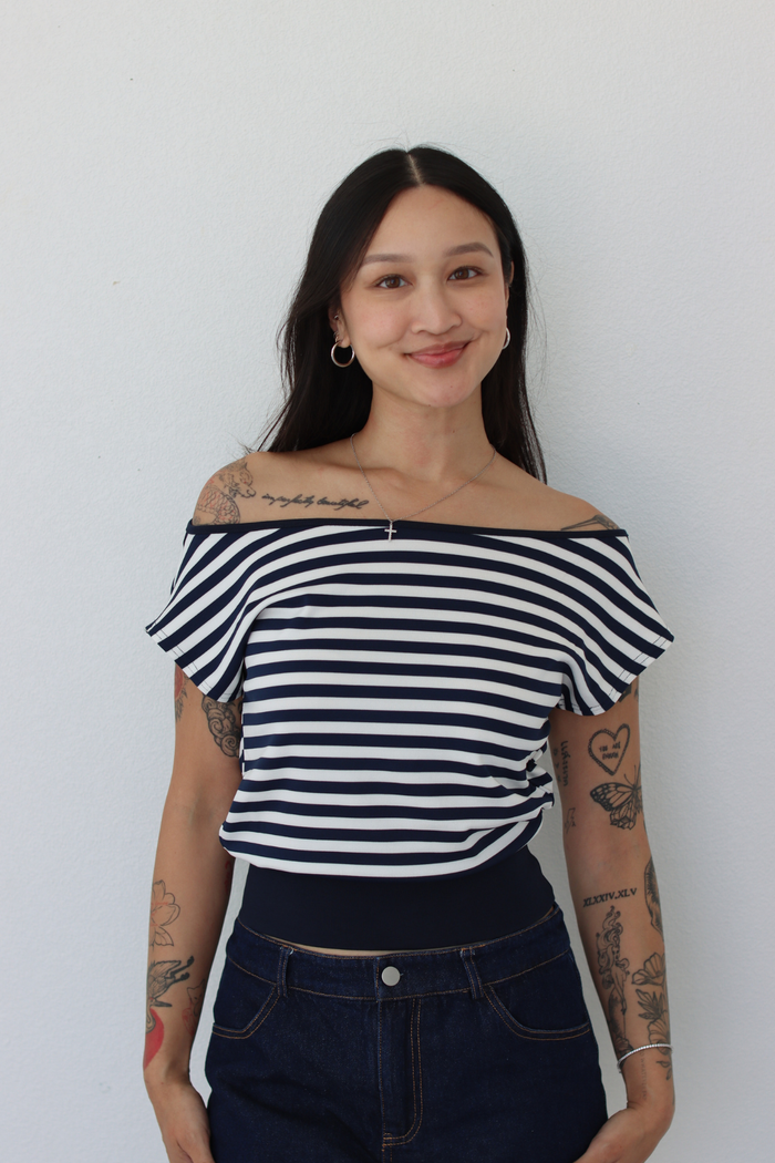 A woman with tattoos wearing a navy and white horizontally striped off-the-shoulder top and dark blue jeans, standing against a plain white wall.