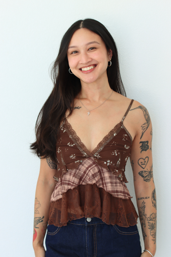 A smiling woman with long dark hair and visible arm tattoos wears a layered brown lace and plaid camisole top with dark blue jeans, standing against a plain light background.