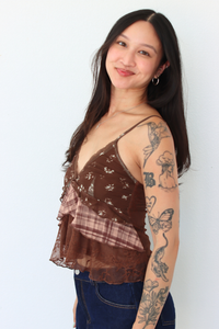 A woman with long dark hair and arm tattoos wearing a brown ruffled camisole top with floral lace and plaid details, paired with blue jeans, standing against a plain white wall.