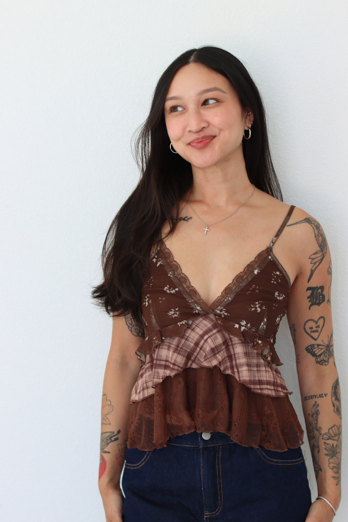 A woman with long dark hair and visible arm tattoos is wearing a brown lace-trimmed camisole top with a ruffled plaid overlay and dark blue jeans, standing against a plain light gray wall.