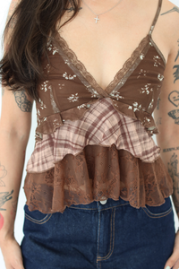 A close-up of a woman wearing a brown ruffled camisole top with floral lace, a plaid panel, and lace trim, paired with blue jeans.